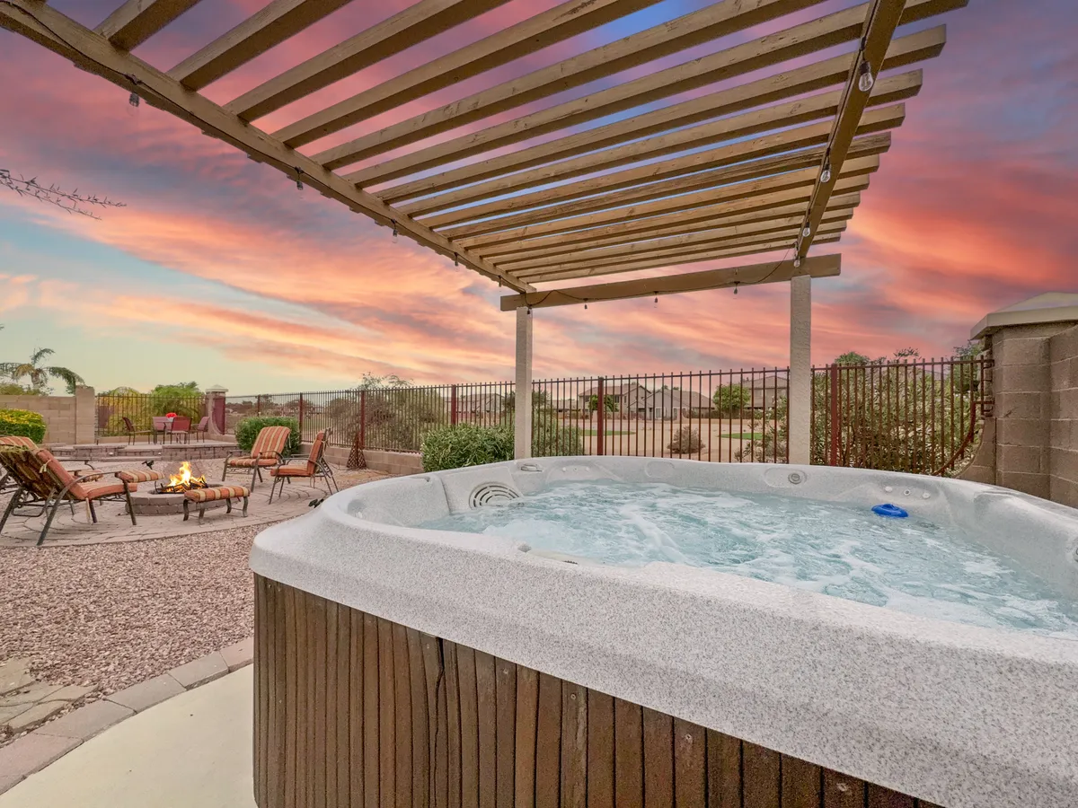 A spacious backyard at sunset featuring a hot tub under a pergola, a modern fire pit with lounge chairs, and a view that stretches toward the golf course beyond. The scene is wrapped in warm golden hues, inviting relaxation and socialization.