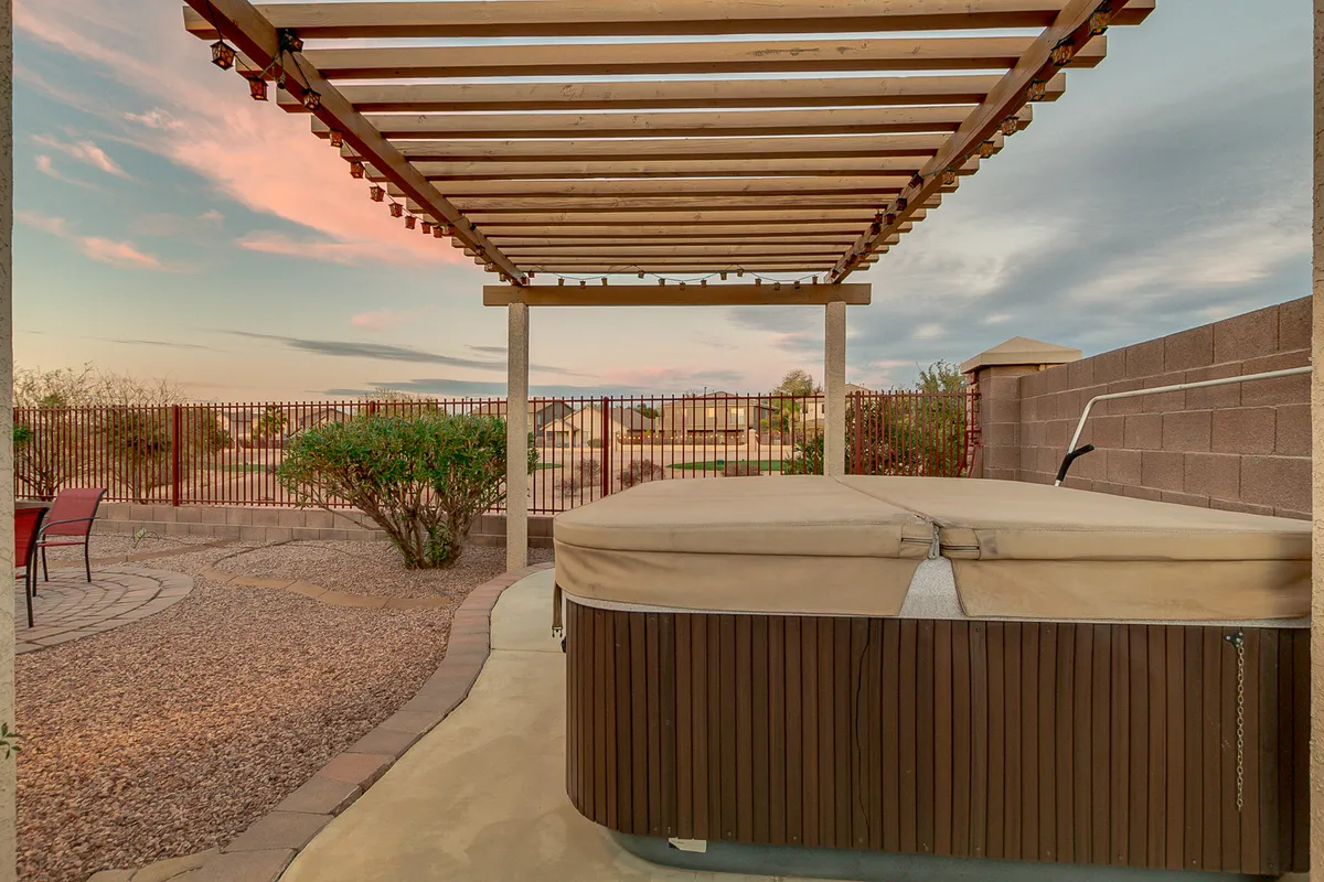 A tranquil backyard setting with a hot tub beneath a pergola, overlooking landscaped grounds. The scene invites outdoor gatherings or quiet moments of reflection, with open skies in the background.