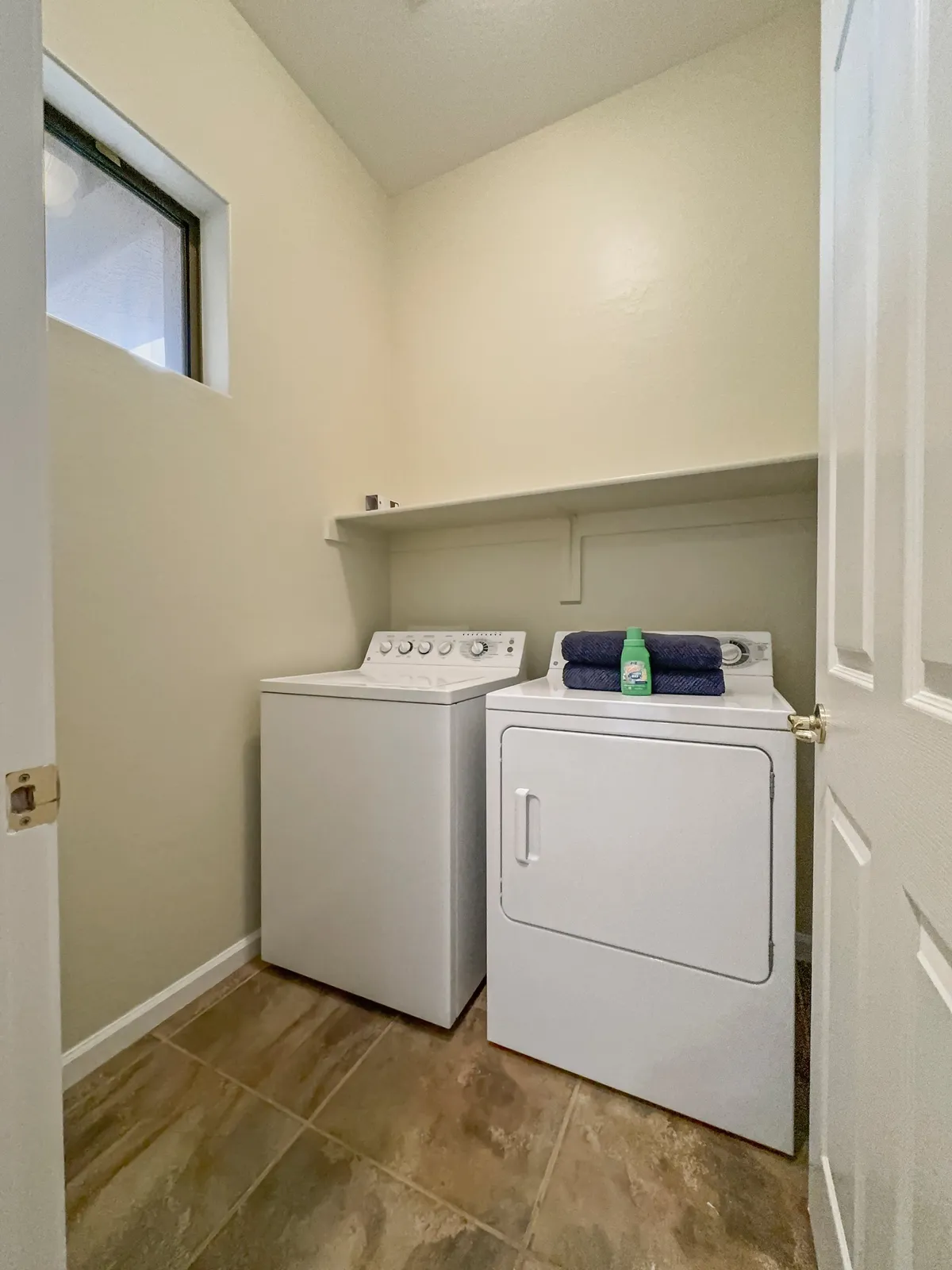 A bright and organized laundry room featuring a modern washing machine and dryer, shelves for storage, and a window that brings in natural light. The space is clean, efficient, and clearly designed for practical daily use.