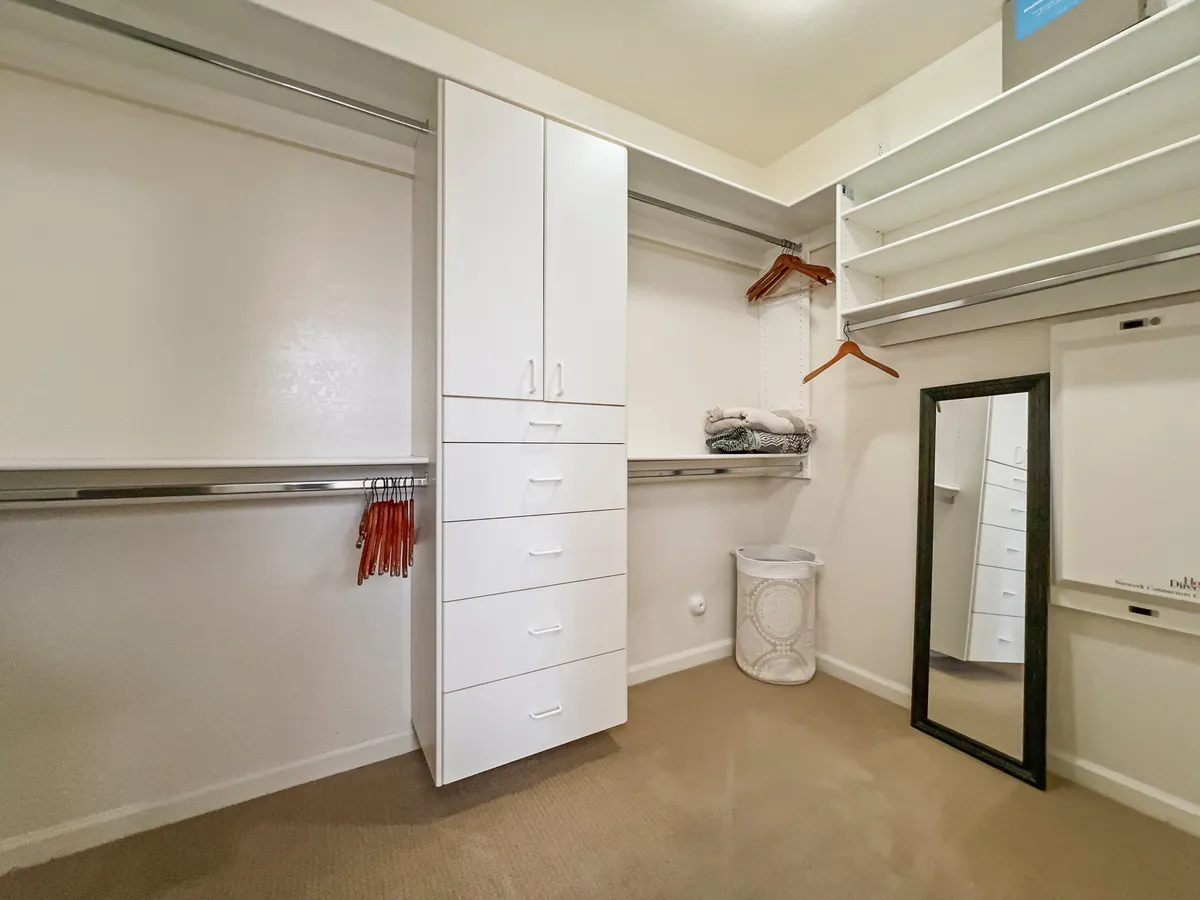 A minimalist interior scene displaying a white storage cabinet, full-length mirror, and open shelving. The area emphasizes orderliness and provides a sense of generous storage options.