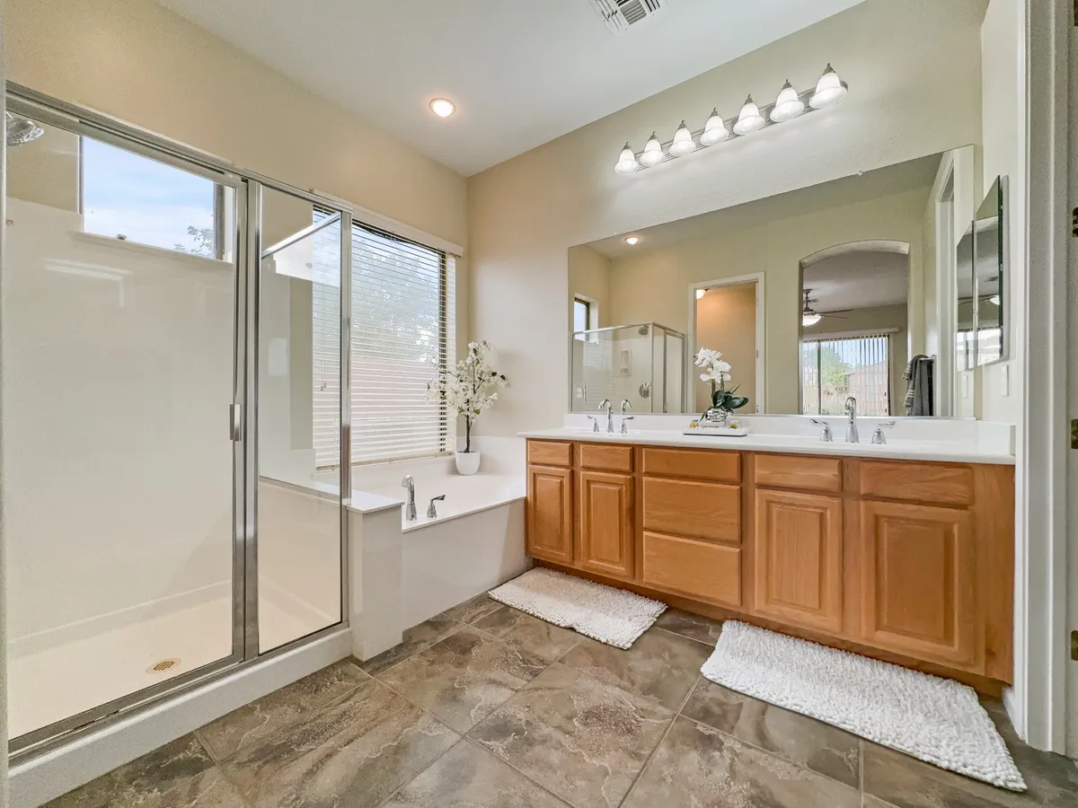 A spacious spa-style bathroom with a double sink vanity, large shower enclosure, and a deep soaking bathtub set next to a sunlit window. A decorative plant adds a touch of greenery atop the vanity, enhancing the room's tranquil, inviting feel.
