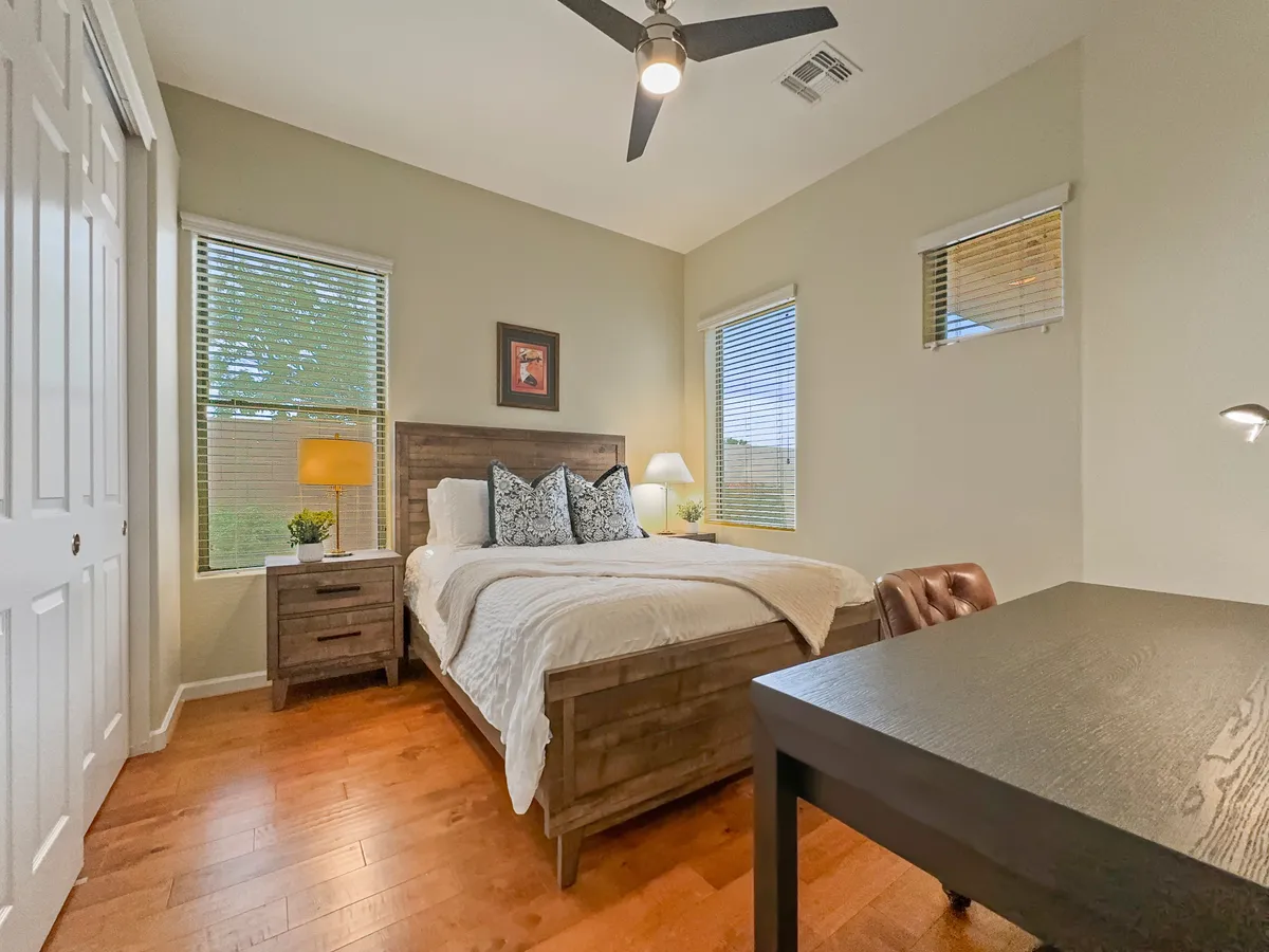 A cozy bedroom-office hybrid featuring a comfortable bed with decorative pillows, a nightstand with a lamp, and a dedicated workspace with a desk and chair. Sunlight streams in, creating an inviting spot for both restful nights and productive work-from-home days.